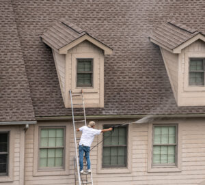 Man standing on a ladder leaning against a house. The man is using a pressure washer to wash the roof of the house.