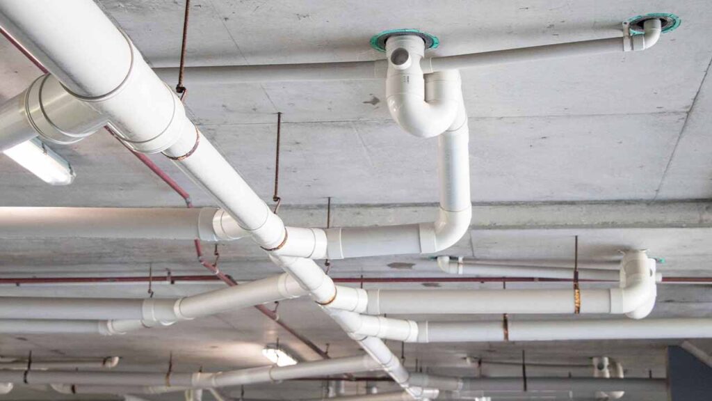 Photo of a commercial car park concrete ceiling. There's a large amount of white PVC pipes protruding from the roof and running in different directions.