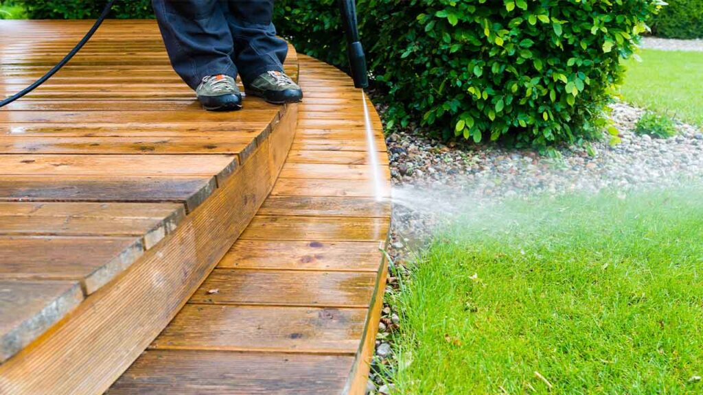 Photo of a person pressure cleaning a wooden deck. All that can be seen is the feet of the person, the pressure cleaning lance spraying water onto the deck