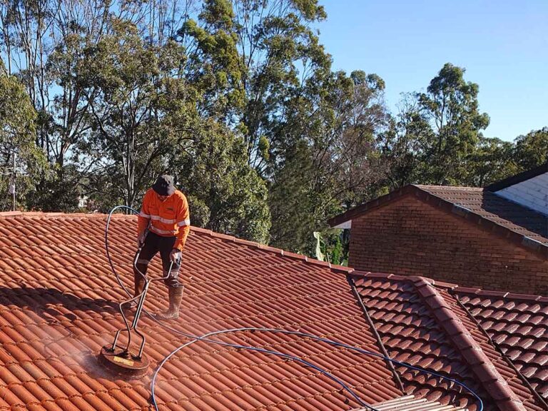 Man on an orange roof using a rotary surface cleaner to pressure wash the roof. There's some tress and another roof visible in the background.