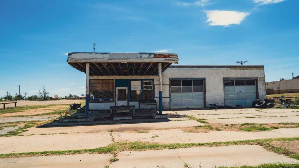 An old abandoned service station. Has 2 pumps and 2 garage doors. The area surrounds the serivce stations has grass growing and dirt. It looks no one has visited in 30 years
