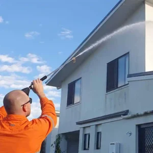 A team member of Pressure washed South East Qld pressure washing the wall of a white two story house. It's a bright day with blue sky, and the water can be seen spraying the wall of the house.