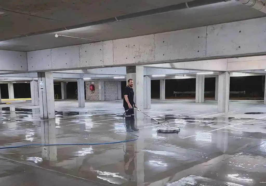 A team member of Pressure Washed South East Qld using a sruface rotary cleaning to clean the concrete floor of a basement car park. The floor is all wet from water cleaning. The car park basement ceiling and pillars are all grey concrete.