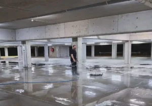 A team member of Pressure Washed South East Qld using a sruface rotary cleaning to clean the concrete floor of a basement car park. The floor is all wet from water cleaning. The car park basement ceiling and pillars are all grey concrete. 