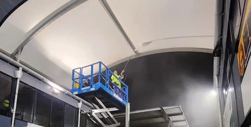 Photo of a team member using a sizzor lift to pressure clean the underneath of a dirty shade sail. It's night time and water mist can be seen in the air from the pressure washer.