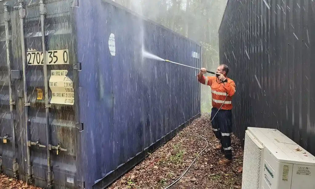 A team member of Pressure Washed South East QLD using a high pressure cleaning gun to high pressure clean a blue colored dirty shipping container. Water can been seen flying everywhere and it's a bright sunny day.