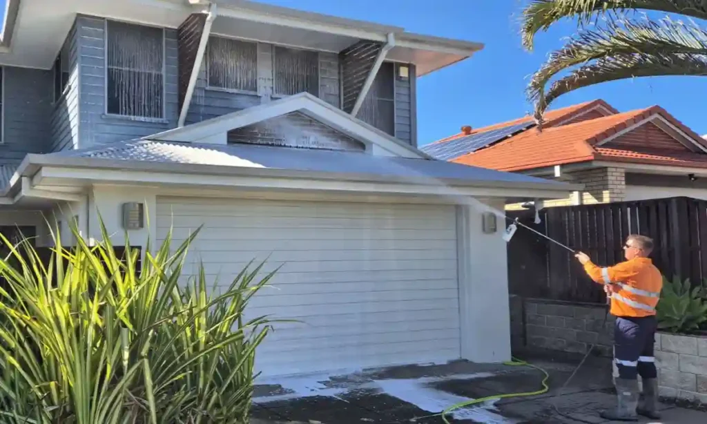 A team member of Pressure Washed South East QLD is standing on the driveway of a 2 story house spraying soap onto the windows and garage door of the house. The front of the house is covered in soap, it's a bright sunny day with blue sky.