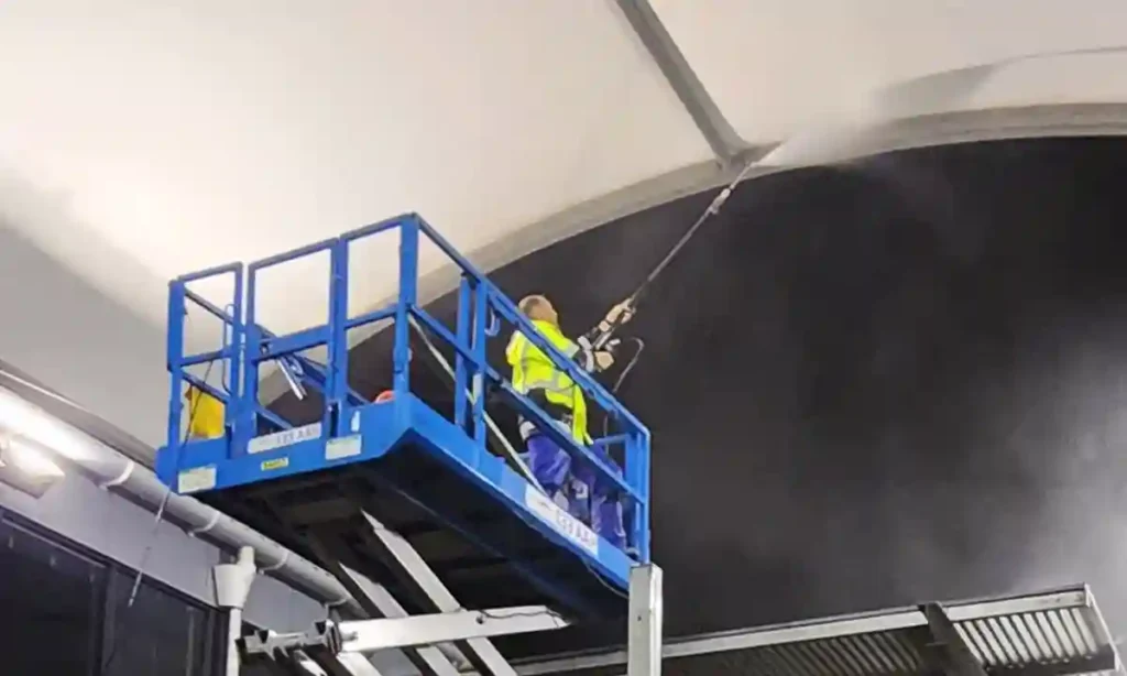 Photo of a team member of Pressure Washed South East QLD pressure cleaning a commercial roof. The cleaner is standing in a scissor lift and is using a long pressure cleaning pole to reach the roof. It's a dark night and water can be seen spraying the roof and reflecting into the dark.